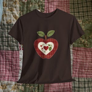 Op de afbeelding: Een bruin t-shirt met een appelontwerp. De appel is rood met een wit hartvormig centrum, versierd met kleine rode bloemen en een groen hart. Groene bladeren en een bruine steel maken het appelontwerp compleet. Het shirt is gemaakt van zacht materiaal.