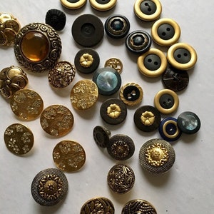 May include: A collection of assorted round buttons in various sizes and designs. The assortment includes metallic gold-tone buttons, matte black buttons, buttons with embedded clear crystals, and buttons featuring intricate embossed floral patterns.