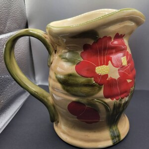 May include: A ceramic pitcher with a floral design. The pitcher is beige with green leaves and red flowers. The pitcher has a green handle.
