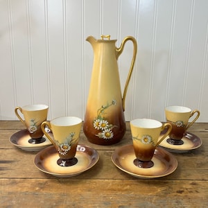 May include: A vintage tea set with a cream-colored teapot and four matching cups and saucers. The set features a delicate floral design with white daisies on a brown background.