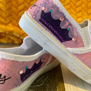 Puede incluir: Un par de zapatos de lona rosa con un dise&ntilde;o de corona morada y pedrer&iacute;a. Los zapatos est&aacute;n decorados con una corona morada y pedrer&iacute;a.