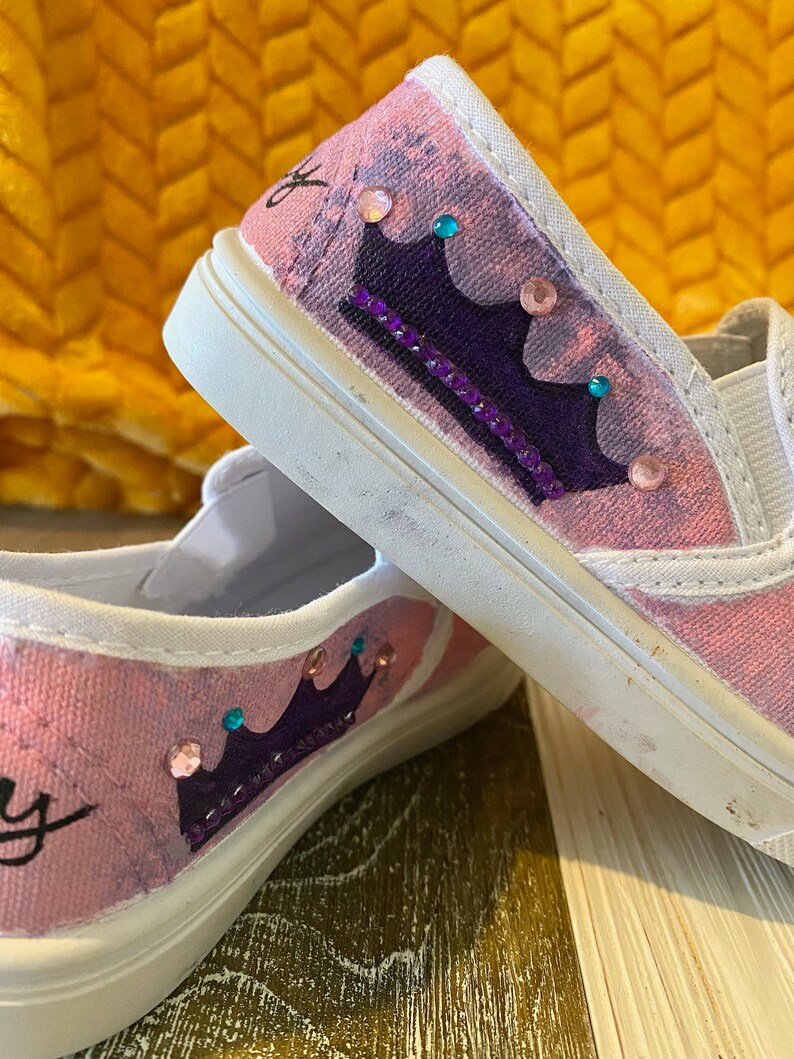 Puede incluir: Un par de zapatos de lona rosa con un dise&ntilde;o de corona morada y pedrer&iacute;a. Los zapatos est&aacute;n decorados con una corona morada y pedrer&iacute;a.