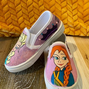 Puede incluir: Un par de zapatos de lona blancos con im&aacute;genes pintadas a mano de Elsa y Anna de la pel&iacute;cula Frozen. Los zapatos tienen un fondo rosa y est&aacute;n decorados con purpurina.
