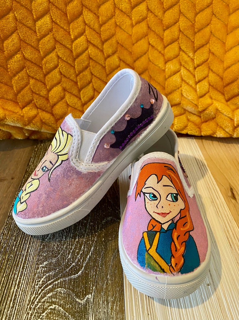 Puede incluir: Un par de zapatos de lona blancos con im&aacute;genes pintadas a mano de Elsa y Anna de la pel&iacute;cula Frozen. Los zapatos tienen un fondo rosa y est&aacute;n decorados con purpurina.