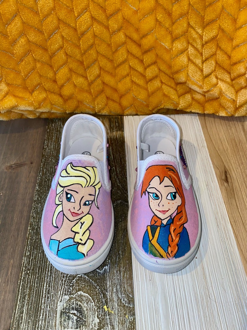 Puede incluir: Un par de zapatos de lona rosa con im&aacute;genes pintadas a mano de Elsa y Anna de Frozen. Los zapatos tienen suelas blancas y cordones blancos.