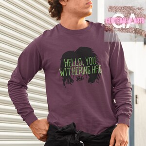 May include: A long-sleeved shirt with a purple background and a black silhouette of a woman's hair with the text "HELLO, YOU WITHERING HAG" in green.