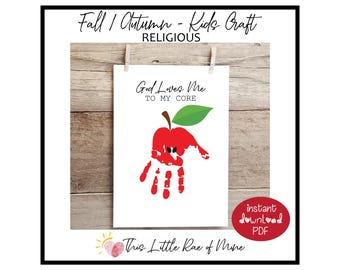May include: A white print with a red handprint apple design, green leaf, and the text "God Loves Me To My Core." The print is clipped to a wooden background with the text "Fall / Autumn - Kids Craft - Religious."