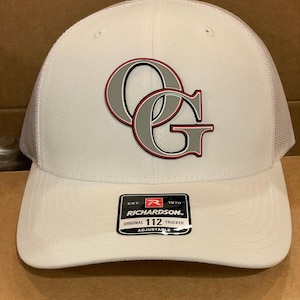 Könnte beinhalten: Weißer Trucker-Cap mit gebogenem Schirm und Mesh-Rückseite. Die Kappe hat ein graues und rotes "OG"-Logo auf der Vorderseite. Ein schwarz-weißes Etikett auf dem Schirm lautet "RICHARDSON ORIGINAL 112 TRUCKER ADJUSTABLE".