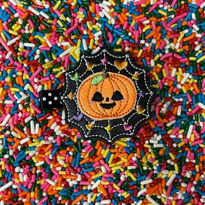 May include: A black hair clip with a Halloween-themed design. The clip features an embroidered orange jack-o'-lantern with a smiling face, surrounded by a black spiderweb design and colorful accents. The background is a vibrant mix of sprinkles.