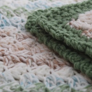 May include: A close-up of a crocheted blanket with a green and white pattern. The blanket is made of soft yarn and has a textured surface.