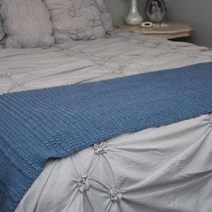 May include: A blue knitted throw blanket on a gray bed with a white ruffled bedspread.