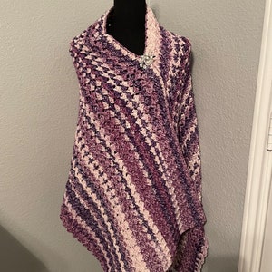 Op de afbeelding: Een handgemaakte gehaakte sjaal in tinten paars en wit, gedrapeerd over een mannequin. De sjaal heeft een gestructureerd steekpatroon en is vastgezet met een decoratieve broche. Het kleurenpalet omvat verschillende tinten paars en wit.