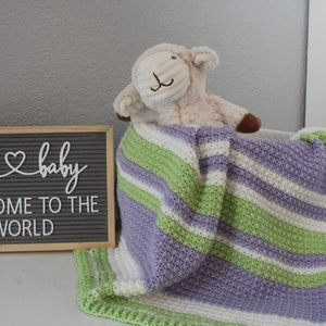 May include: A purple, green, and white striped crocheted baby blanket with a plush white lamb toy on top. The blanket is on a white surface with a letter board that says "oh baby welcome to the world" in the background.