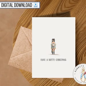 Op de afbeelding: Een witte kaart met een cartoon-notenkrakerillustratie en de tekst "Have a Nutty Christmas". De kaart ligt op een houten oppervlak met een bruine envelop en een logo voor "Rabbits Quill Personalized Greeting Cards".