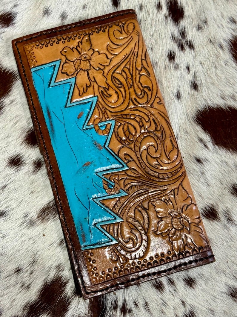 Peut inclure: Un portefeuille en cuir fait &agrave; la main avec un motif en zigzag turquoise sur le c&ocirc;t&eacute; gauche. Le portefeuille pr&eacute;sente des sculptures florales et en volutes complexes dans des tons de beige et de marron. Les bords sont marron fonc&eacute;.