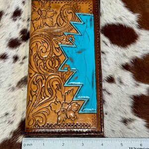 Peut inclure: Un portefeuille en cuir marron avec des motifs floraux et des zigzags turquoise. Le portefeuille mesure environ 15 cm de long et pr&eacute;sente des motifs en relief complexes. La section turquoise ajoute une touche de couleur.