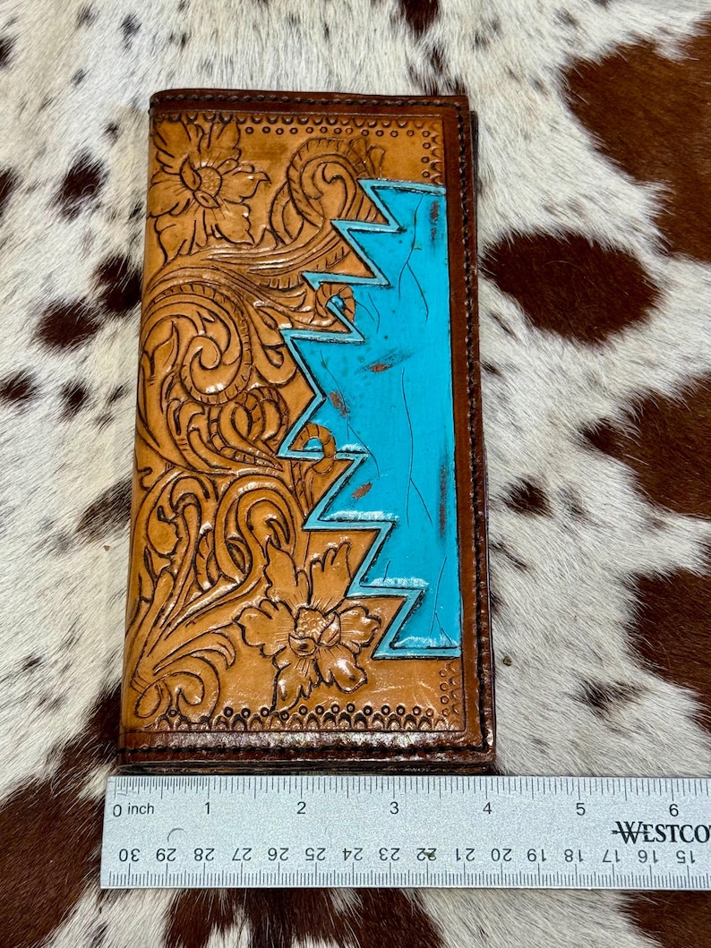 Peut inclure: Un portefeuille en cuir marron avec des motifs floraux et des zigzags turquoise. Le portefeuille mesure environ 15 cm de long et pr&eacute;sente des motifs en relief complexes. La section turquoise ajoute une touche de couleur.