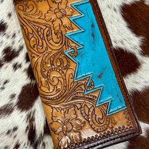 Peut inclure: Un portefeuille en cuir fait &agrave; la main avec des motifs floraux complexes et un motif en zigzag turquoise. Le portefeuille est de couleur marron clair avec des bordures marron fonc&eacute; et un motif floral en relief d&eacute;taill&eacute;.