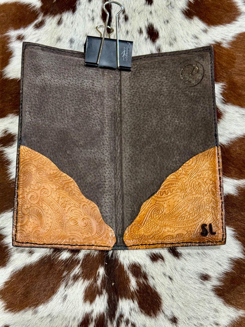 Peut inclure: Portefeuille en cuir marron ouvert avec des accents de design paisley tan en relief. Le portefeuille est maintenu ouvert avec une pince &agrave; reliure noire. Le fond est un motif de peau de vache.