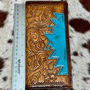 Peut inclure: Un portefeuille en cuir marron avec des motifs floraux et g&eacute;om&eacute;triques. Le portefeuille pr&eacute;sente une section bleu turquoise et mesure environ 20 cm de long. Le travail de repoussage et le contraste des couleurs cr&eacute;ent un look unique et artisanal.