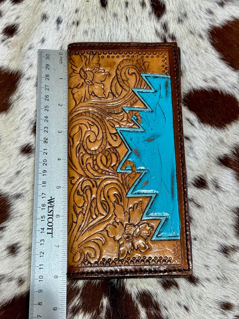 Peut inclure: Un portefeuille en cuir marron avec des motifs floraux et g&eacute;om&eacute;triques. Le portefeuille pr&eacute;sente une section bleu turquoise et mesure environ 20 cm de long. Le travail de repoussage et le contraste des couleurs cr&eacute;ent un look unique et artisanal.