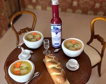 May include: A table setting with three bowls of chicken noodle soup, a challah bread, and a bottle of Manischewitz Concord Grape wine. The table is set with white plates, silverware, and glasses.