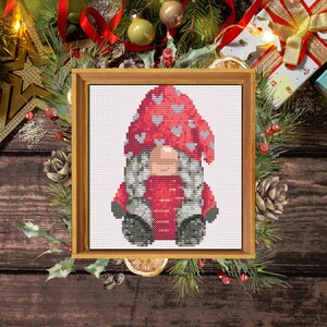 Può includere: Uno schema di punto croce di un gnomo che indossa un cappello rosso con cuori bianchi e un cappotto rosso. Lo gnomo è circondato da una cornice di legno.