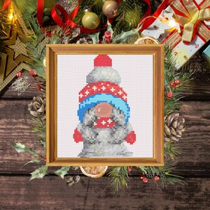May include: Cross stitch pattern of a gnome wearing a red and white hat with a blue scarf and red mittens. The gnome is standing in front of a white background.