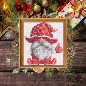 Könnte beinhalten: Ein roter und weißer Zwerg mit weißem Bart und roter Mütze ist in einem Kreuzstichmuster dargestellt. Der Zwerg steht mit ausgestreckten Armen da, als würde er winken. Der Kreuzstich ist in einem Holzrahmen gerahmt.