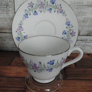 May include: A white teacup and saucer set with a floral design. The cup and saucer feature a delicate blue and purple floral pattern around the rim. The saucer has a gold trim.