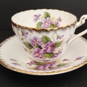 May include: A white teacup and saucer with a floral pattern of purple violets and green leaves. The cup and saucer have a gold trim around the edges.