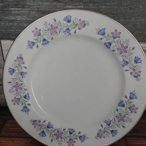 May include: White ceramic plate with a silver rim and a floral design. The design features blue and purple bellflowers and green leaves arranged in a garland around the plate's edge. The plate is displayed on a clear stand.