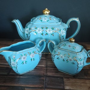 May include: A blue teapot, creamer, and sugar bowl set with gold trim and floral designs. The teapot has a lid with a gold knob. The creamer and sugar bowl have matching floral designs.