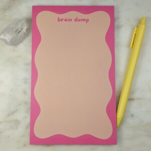 May include: A pink notepad with a wavy edge and the words "brain dump". A yellow pen lies on the right. A clear crystal is on the left. The notepad is for jotting down ideas. The notepad's border is a vibrant pink.