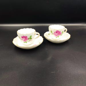 Può includere: Due tazze da tè bianche e piattini con un motivo di rosa rosa. Le tazze e i piattini hanno un bordo dorato.