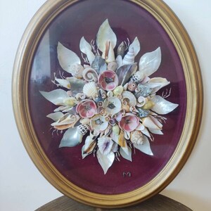 May include: A framed seashell artwork with a burgundy background. The seashells are arranged in a flower shape with pink and white accents. The frame is gold and oval shaped. The piece is signed with the letter "M".