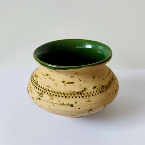 Puede incluir: Una pequeña maceta redonda de cerámica con un borde verde esmaltado y un cuerpo beige. La maceta tiene una superficie texturizada con un patrón de pequeños puntos y líneas.