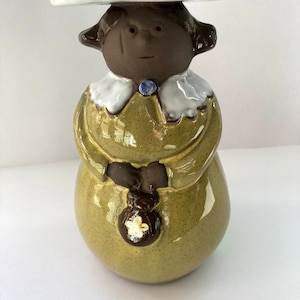 May include: A ceramic figurine of a person wearing a hat with blue and green details. The figurine has a yellow-green dress, a white collar, and a small blue brooch. A small brown purse with a white flower is held in the hands.