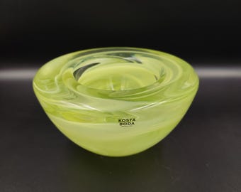 May include: A green glass candle holder with a swirling design. The holder has the text "Kosta Boda" on the bottom.
