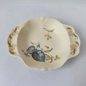 May include: A white oval serving dish with a floral design in shades of blue, green, and gold. The dish has a scalloped edge and two handles.