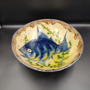 Può includere: Una ciotola in ceramica con un design di pesce blu e giallo. La ciotola ha un bordo marrone e uno sfondo bianco. Il pesce è circondato da alghe verdi.