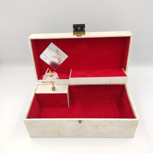 May include: A white music box with a ballerina figurine inside. The box has a red velvet interior and a small mirror on the lid.