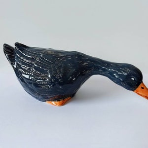 May include: A ceramic goose figurine with a dark blue glaze and orange beak. The goose is in a standing position with its head bent down.
