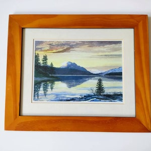 May include: A framed watercolour painting depicting a mountain lake scene with a sunset sky, trees, and a mountain range reflected in the water.