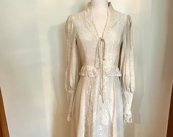 May include: A vintage cream-colored dress with long sleeves and a high collar. The dress features intricate lace detailing on the bodice, sleeves, and skirt. A tie closure accents the neckline. The dress has a ruffled hem and cuffs, and is displayed on a mannequin.