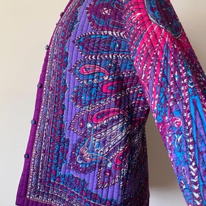 May include: A quilted jacket featuring a vibrant paisley pattern in shades of purple, magenta, and bright blue. The garment has a vertical quilted texture, a round neckline, and a front closure with small, matching fabric-covered buttons.