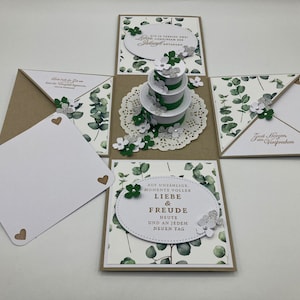 Könnte beinhalten: Eine weiße und grüne Explosionsbox mit einem dreistöckigen Kuchen-Design. Die Box hat einen weißen Hintergrund mit grünen Eukalyptusblättern und weißen Blüten. Der Text auf der Box lautet "Auf unzählige Momente voller Liebe & Freude Heute und an jedem neuen Tag" und "Zwei Herzen ein Versprechen".