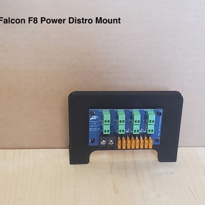 May include: A black plastic mount for a Falcon F8 Power Distro. The mount has a cutout for the power distro and a label that reads "Power Distro 12V 10A".