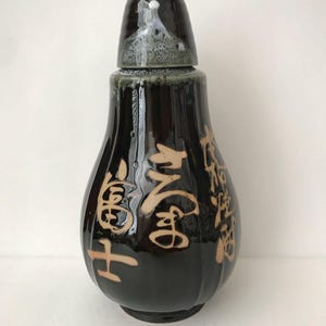 May include: A dark brown ceramic bottle with a matching lid, featuring a glossy finish and light-colored Japanese calligraphy characters etched into the surface. The bottle has a bulbous, pear-shaped body with vertical ridges and a tapered neck.
