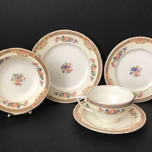 May include: A cream-colored tea set with floral and gold trim. The set includes a teacup and saucer, two plates, and a bowl. The floral design features pink, blue, and yellow flowers. The set is displayed on a black surface.